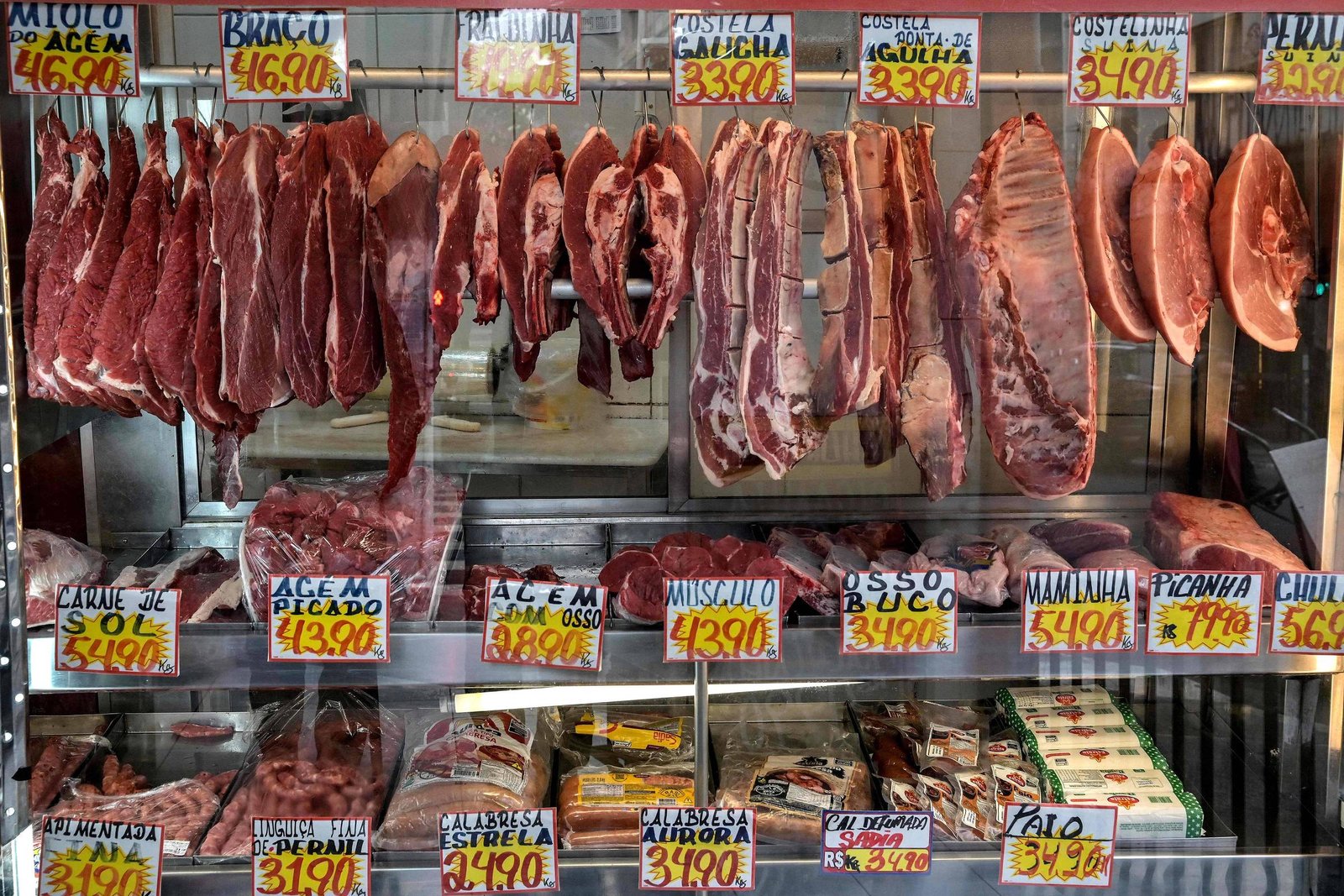 Diversos cortes de carne bovina pendurados em ganchos e embalados em bandejas sobre balcão. Etiquetas amarelas indicam preços variados por quilo, com destaque para picanha, costela e alcatra.