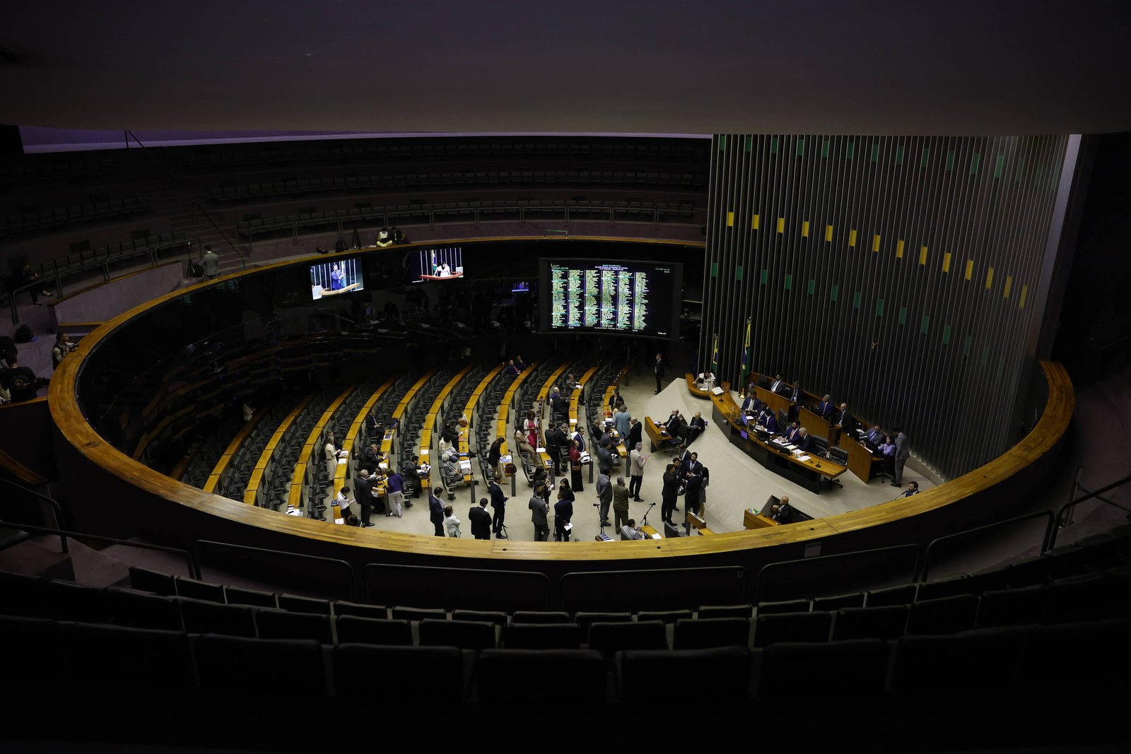 Plenário de câmara legislativa com assentos em semicírculo ocupados por parlamentares e pessoas em pé no centro. Painéis eletrônicos exibem informações ao fundo, e mesa diretora está à direita.