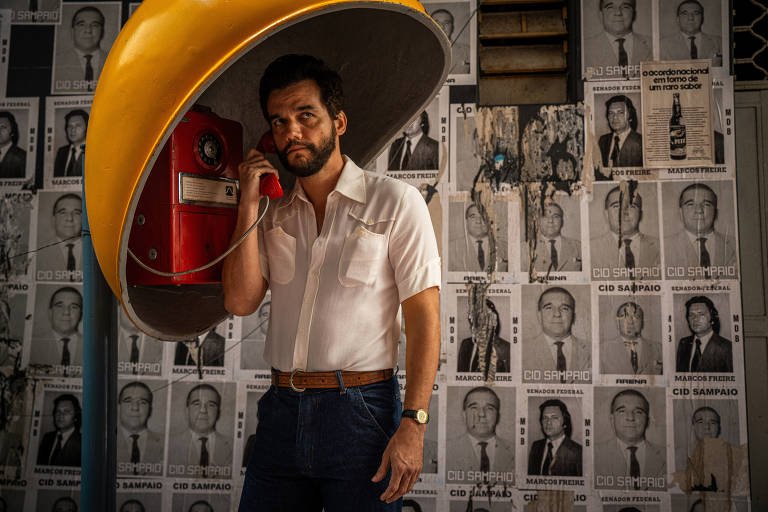 Homem de barba e cabelo escuro usa telefone público amarelo com cabine vermelha. Ele veste camisa branca e calça jeans, está em pé diante de parede coberta por cartazes pretos e brancos com fotos e textos.