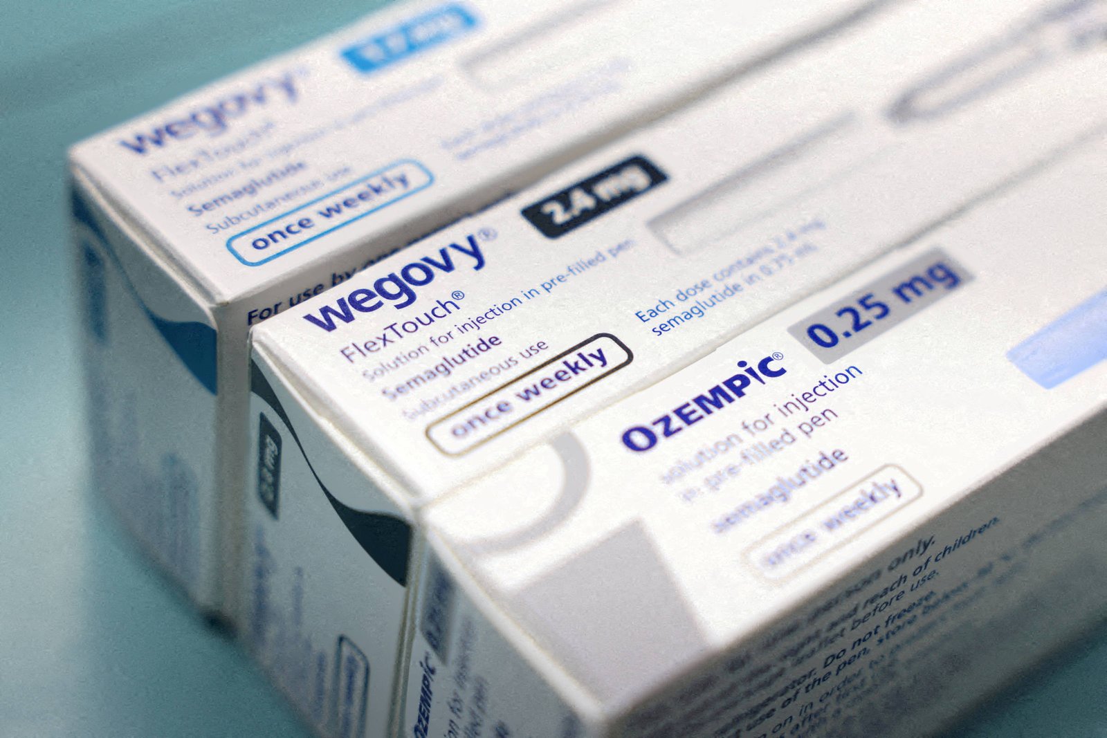 Caixas brancas de medicamentos Wegovy e Ozempic alinhadas sobre superfície azul. Ambas indicam uso semanal e dosagens visíveis: Wegovy 2,4 mg e Ozempic 0,25 mg.