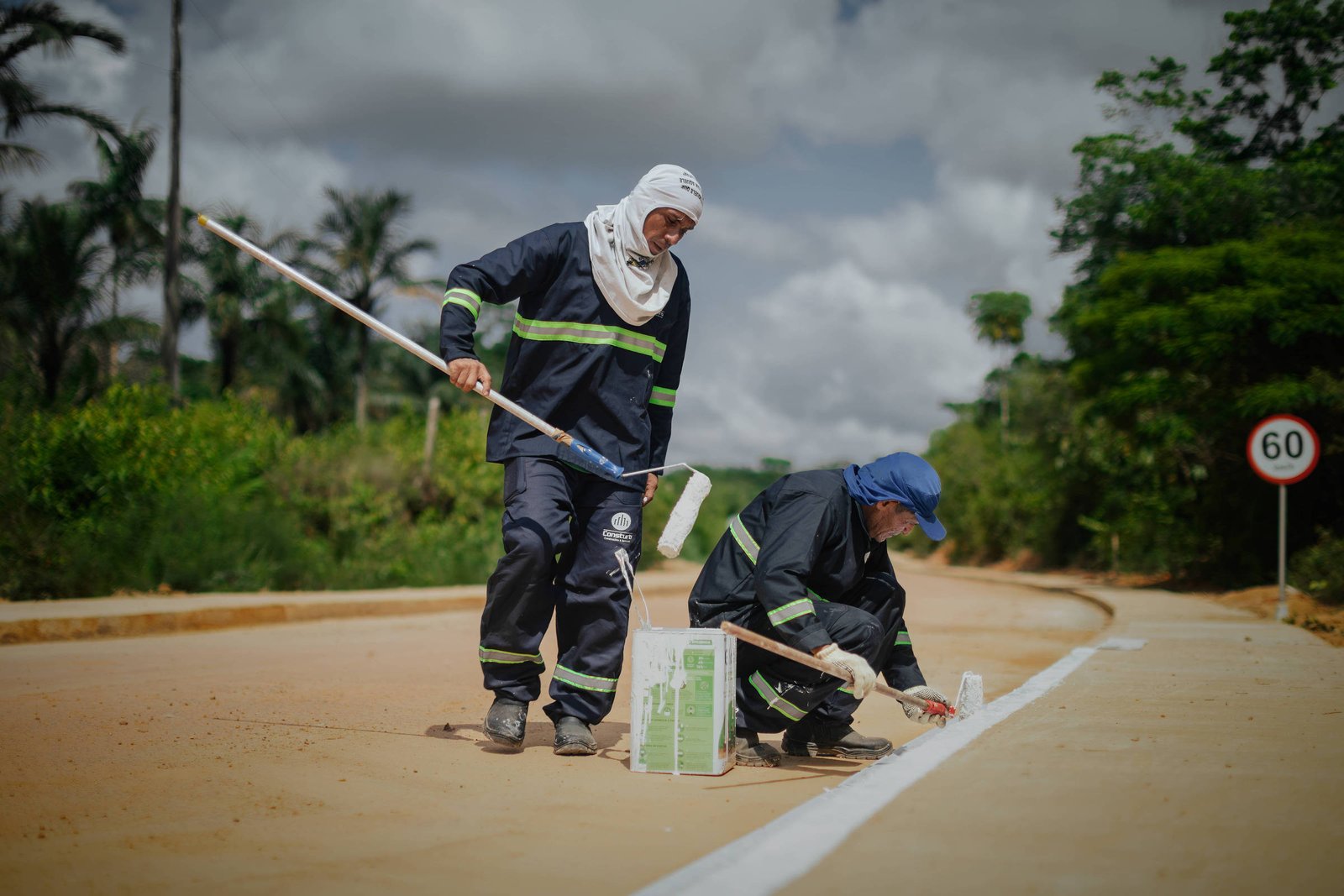 Dois trabalhadores com roupas de proteção pintam uma faixa branca no meio de uma estrada de terra. Um está agachado aplicando tinta com pincel, o outro está em pé segurando um rolo de pintura. Ao fundo, vegetação densa e placa de limite de velocidade 60 km/h.