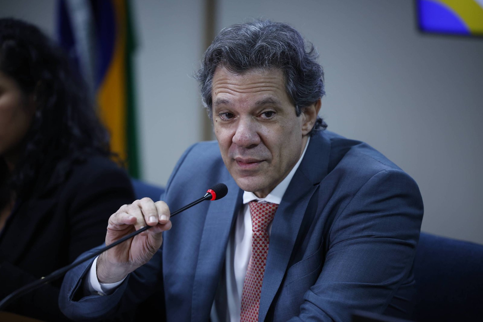 Homem de meia-idade com cabelo grisalho veste terno azul e gravata vermelha, falando em microfone em ambiente formal. Fundo desfocado mostra bandeira e outra pessoa ao lado.