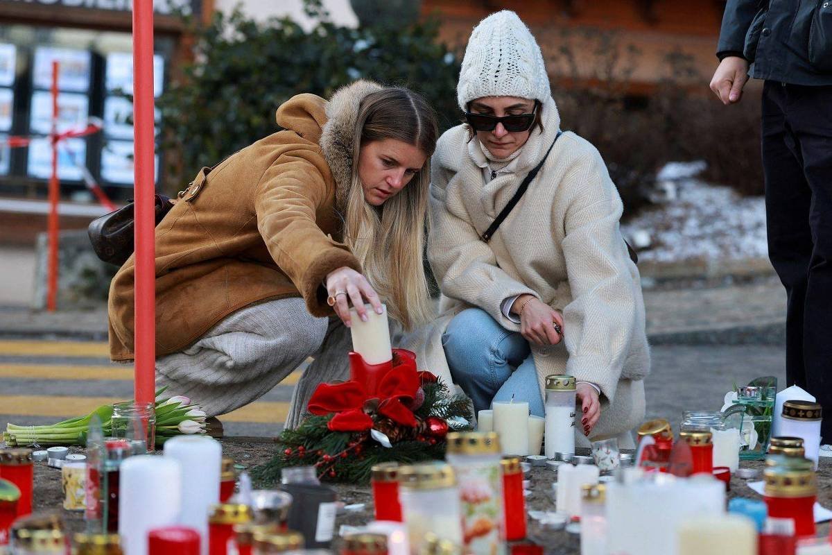 Duas mulheres agachadas acendem velas em um memorial com várias velas e flores vermelhas. Uma veste casaco marrom e a outra casaco branco com gorro e óculos escuros. Ambiente externo com vegetação ao fundo.