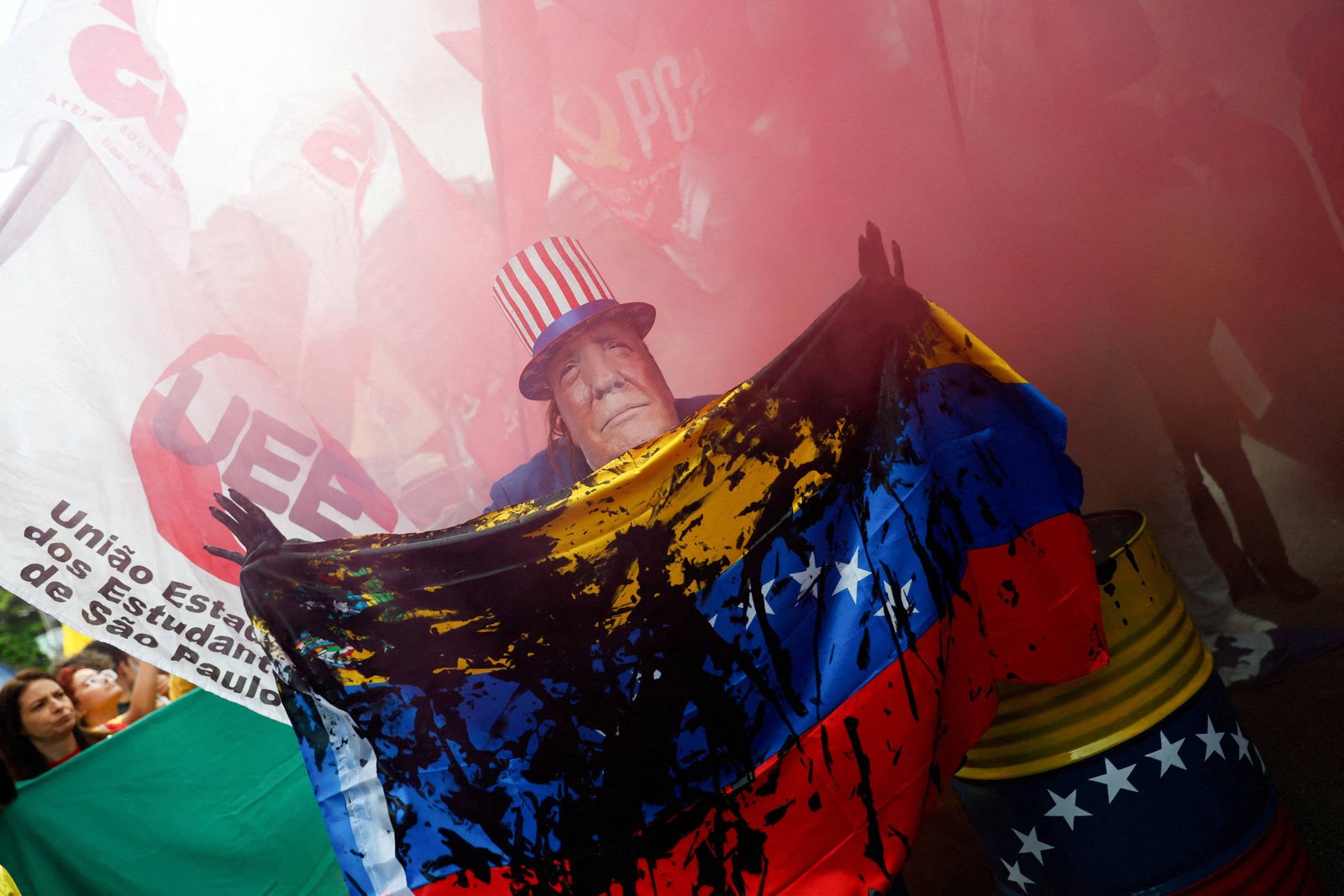 Pessoa com máscara segurando bandeira da Venezuela manchada de tinta preta durante protesto com fumaça vermelha e bandeiras ao fundo.