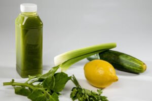 Garrafa plástica com tampa branca contendo suco verde ao lado de um pepino, um talo de aipo, um limão amarelo, folhas verdes e um ramo de salsa sobre superfície branca.