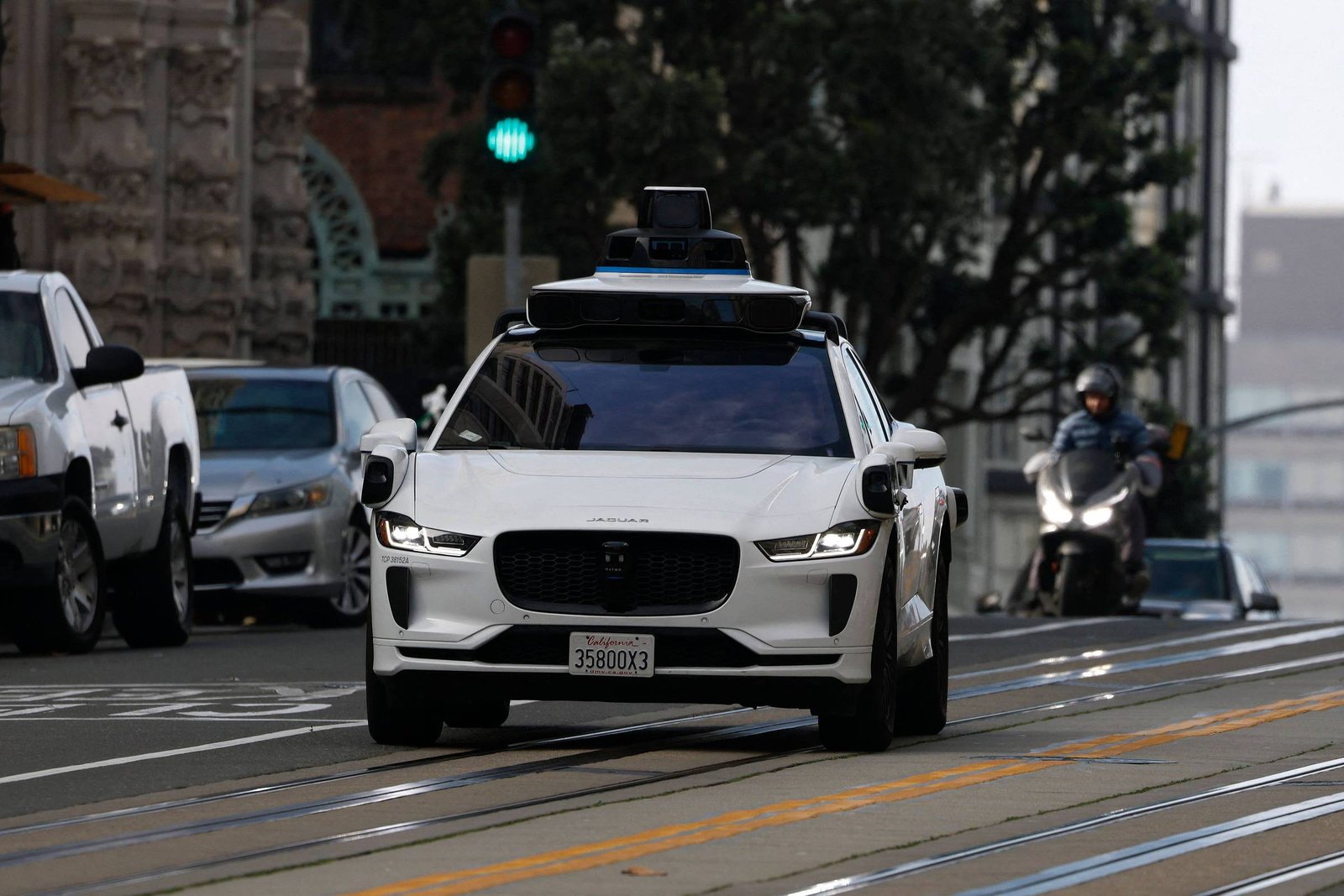 Carro branco autônomo equipado com sensores no teto circula em via urbana com semáforo verde. Veículos e motociclista aparecem ao fundo em ambiente de cidade.