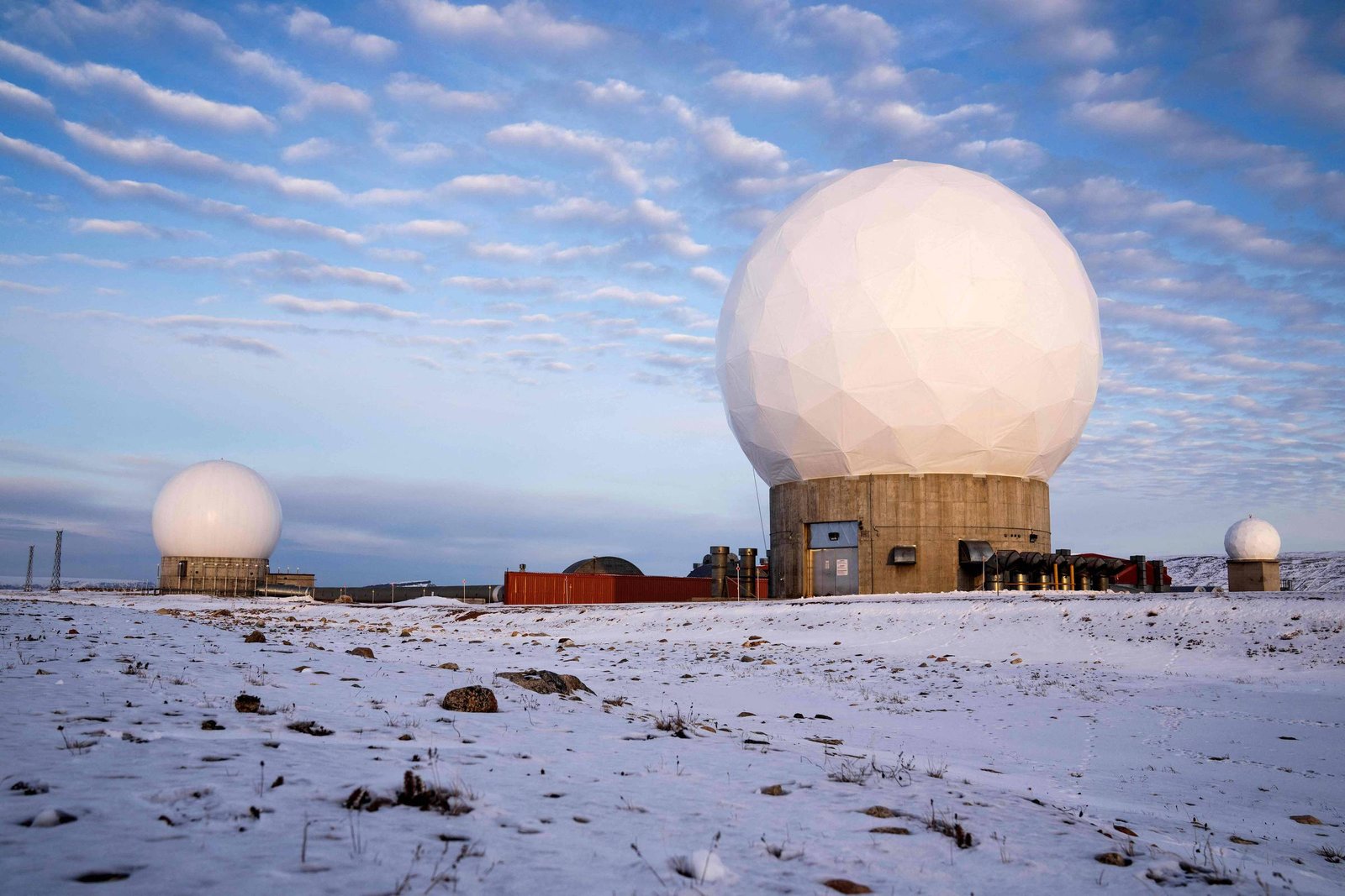Três grandes radares esféricos brancos sobre bases cilíndricas em terreno coberto por neve. Céu azul com nuvens dispersas ao fundo.