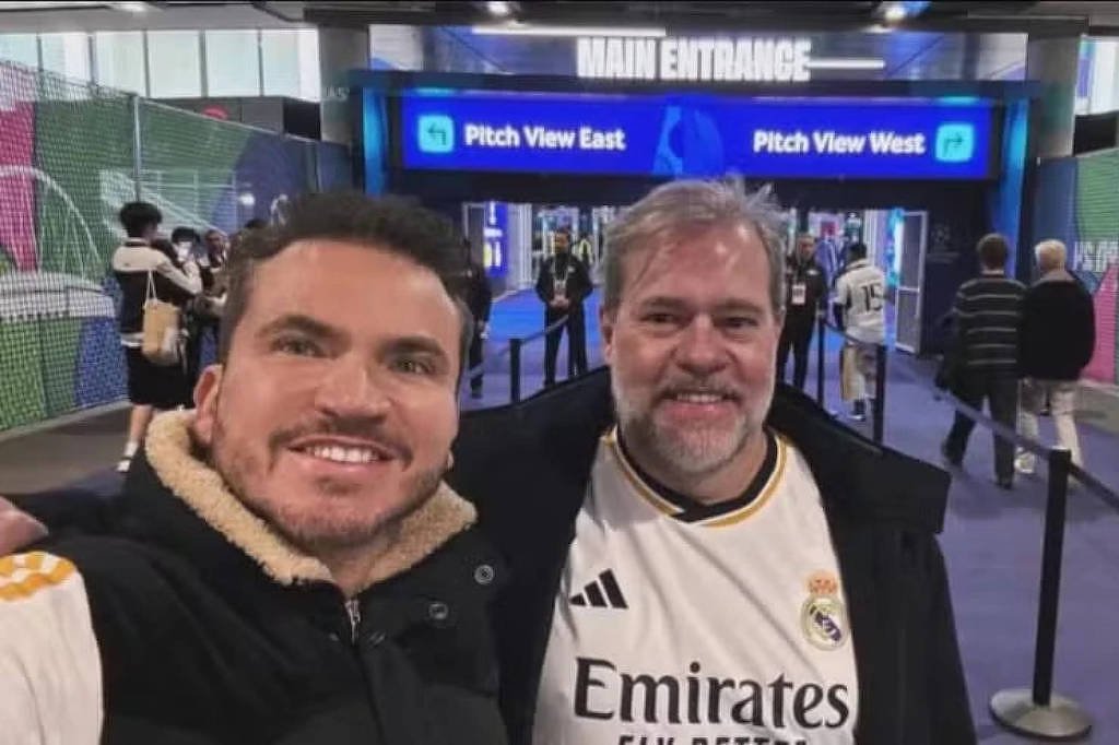 Dois homens sorriem para a câmera na entrada principal de um estádio. Um deles veste jaqueta preta com gola de pelo e o outro camisa branca do Real Madrid. Ao fundo, placas indicam acesso às áreas Pitch View East e Pitch View West.