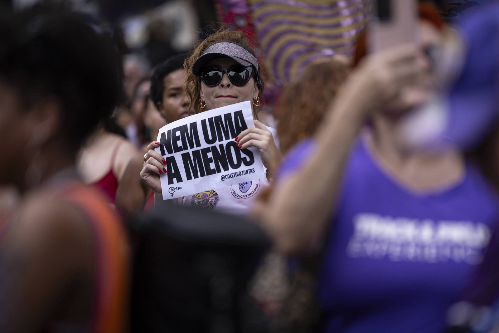 Mulher com óculos escuros e boné segura cartaz com a frase