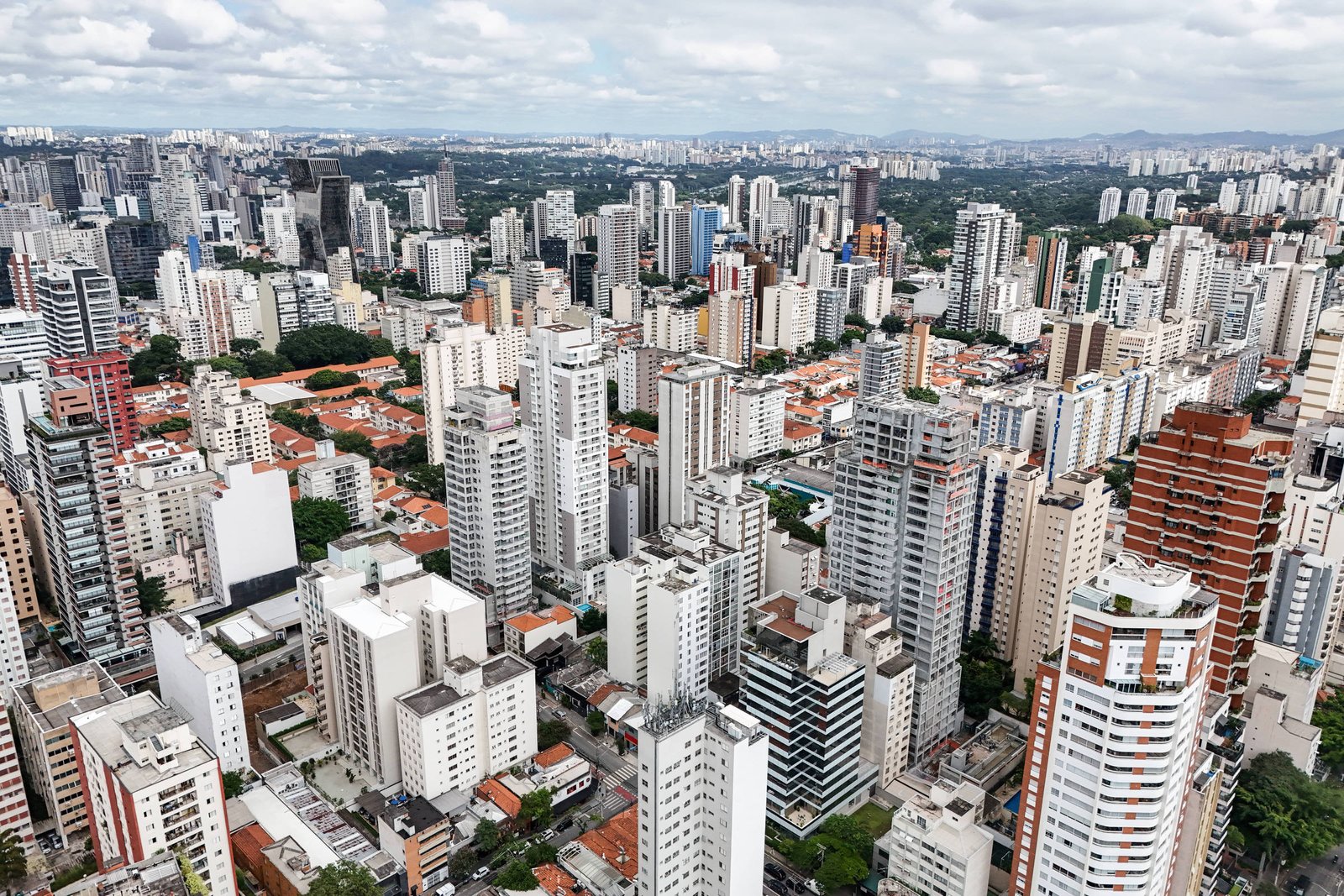 A imagem mostra uma vista aérea de uma cidade com muitos edifícios altos e baixos. Há uma mistura de prédios residenciais e comerciais, com algumas áreas de telhados vermelhos visíveis entre os edifícios. O céu está parcialmente nublado, e ao fundo, é possível ver mais prédios e uma linha de montanhas ao longe.