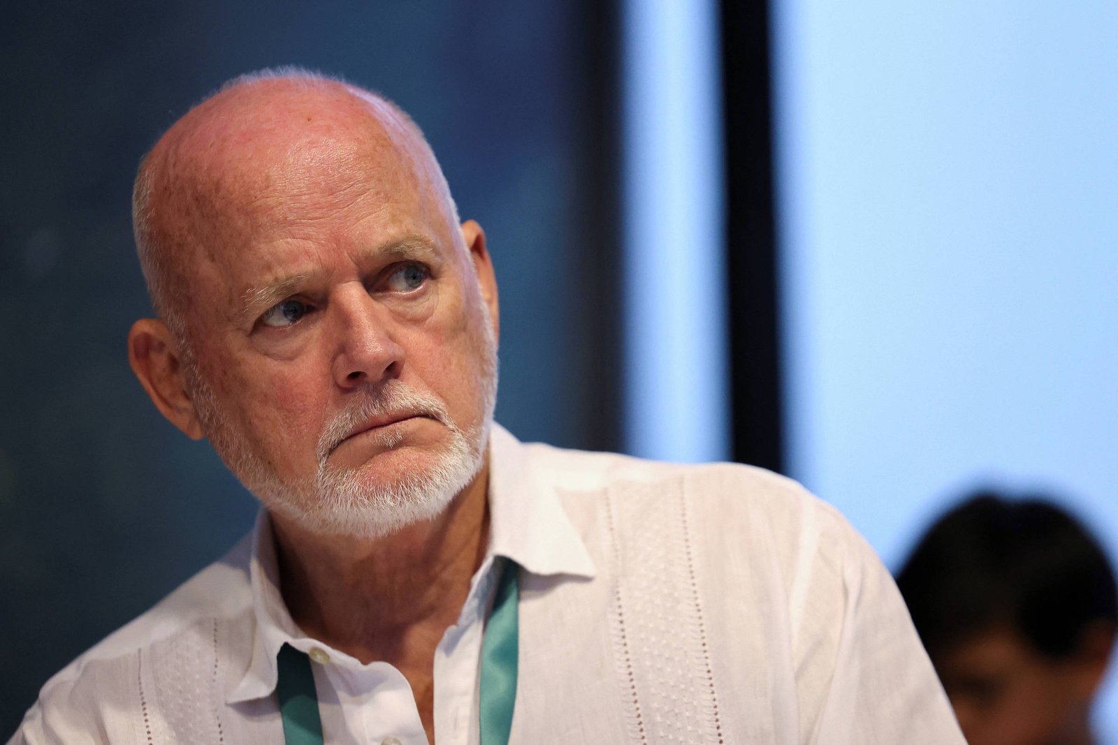 Homem idoso com cabelo ralo e barba branca usa camisa branca e crachá verde, olhando para o lado em ambiente interno com fundo desfocado.