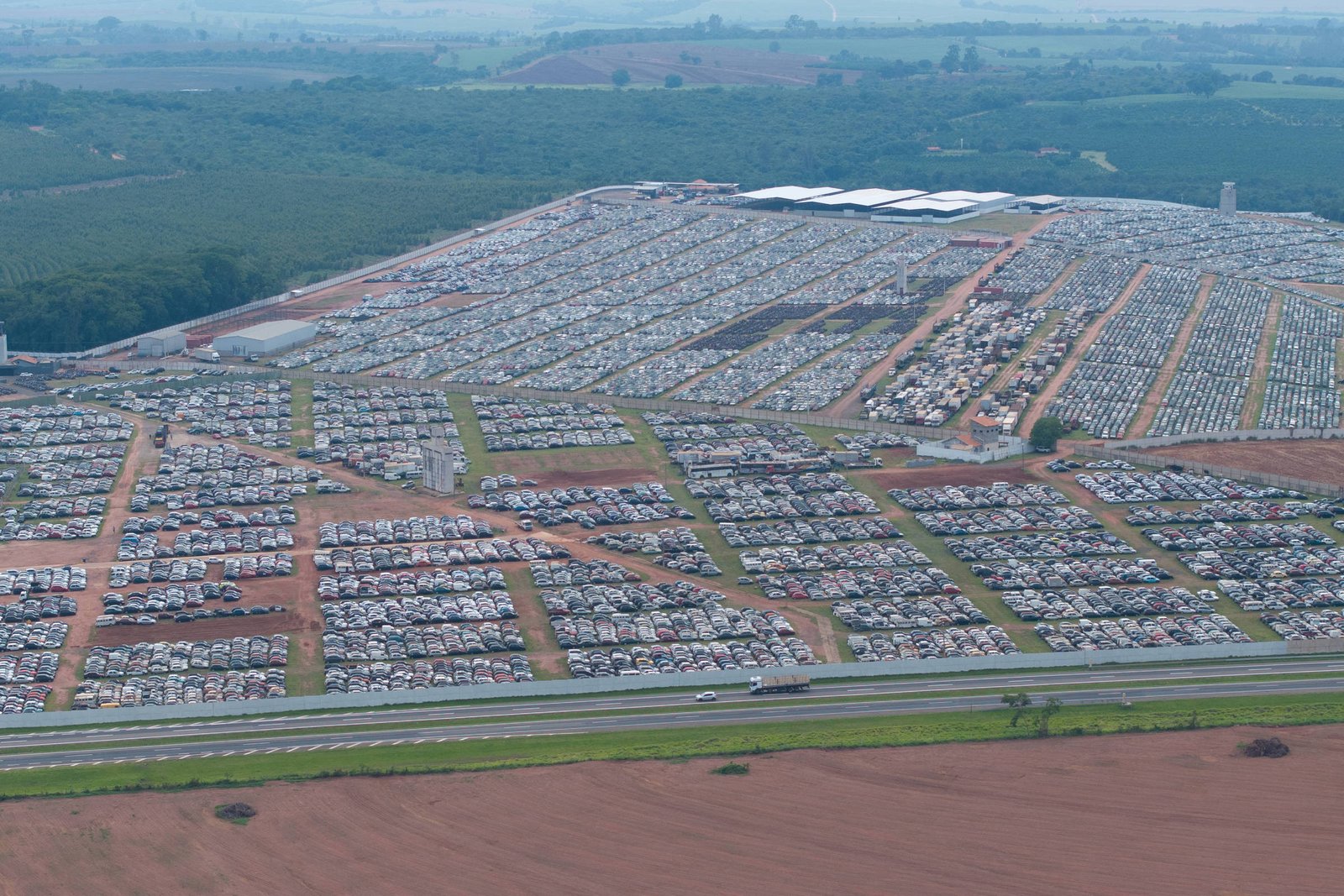 Vista aérea mostra um enorme estacionamento com milhares de carros organizados em lotes separados por vias de terra. O local está cercado por áreas verdes e campos agrícolas ao fundo.