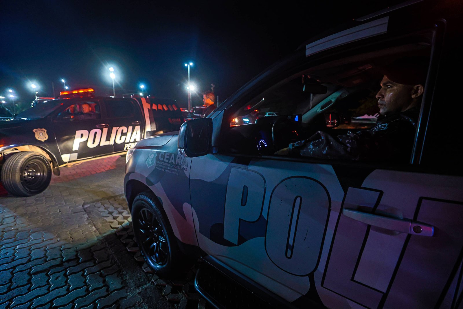 Duas viaturas da polícia estão estacionadas em área pavimentada à noite, com luzes de emergência acesas. Um policial está visível dentro de uma das viaturas, observando a cena.