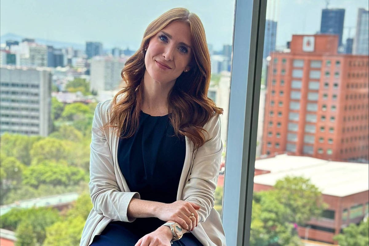 Mulher de cabelos longos e ruivos sentada em banco alto, vestindo calça e blusa azul escuro com blazer branco, em ambiente interno com grande janela de vidro. Ao fundo, vista de prédios, árvores e céu claro em área urbana.