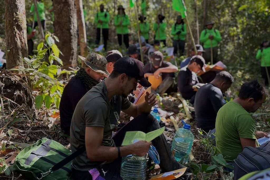 Pessoas sentadas no chão de uma área de mata, algumas lendo documentos ou livros. Ao fundo, grupo em pé veste camisetas verdes e segura bandeiras verdes. Ambiente com árvores e vegetação densa.