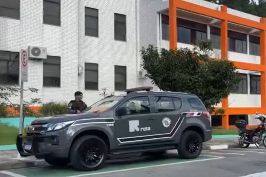 Viatura preta da Polícia Civil estacionada em vaga demarcada em frente a prédio branco com detalhes laranja. Dois policiais estão próximos ao veículo, e há uma motocicleta e outro carro estacionados ao lado.