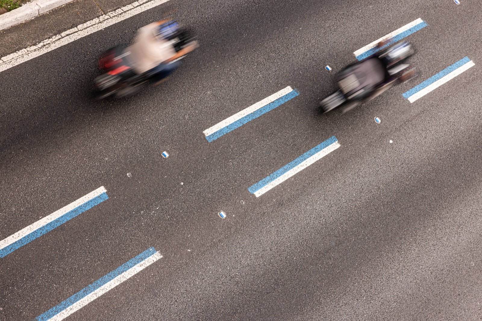 Duas motocicletas em alta velocidade trafegam em pista asfaltada com faixas de sinalização azul e branca. A imagem é capturada de cima, mostrando o asfalto e as linhas de divisão das faixas.