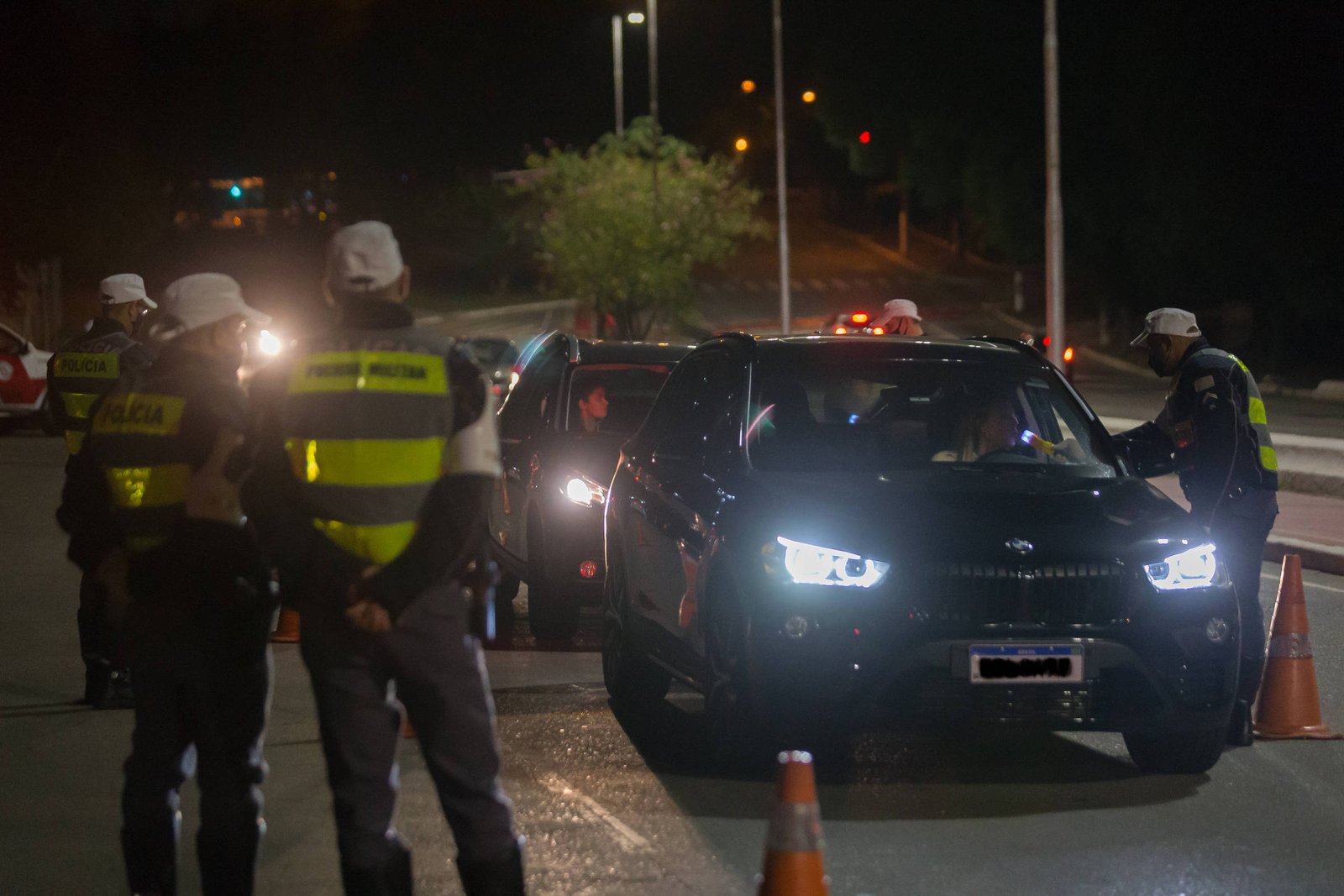 Três policiais em coletes refletores abordam veículo preto parado em blitz noturna. Cone laranja delimita área, postes iluminam local.