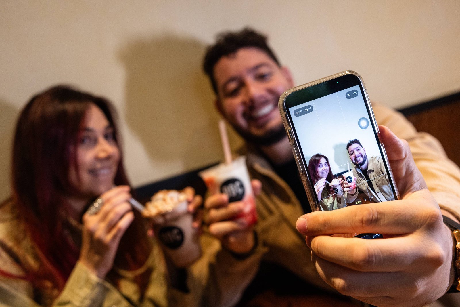 Casal sorridente segura copos de bebidas com canudos enquanto faz selfie com smartphone em ambiente interno.