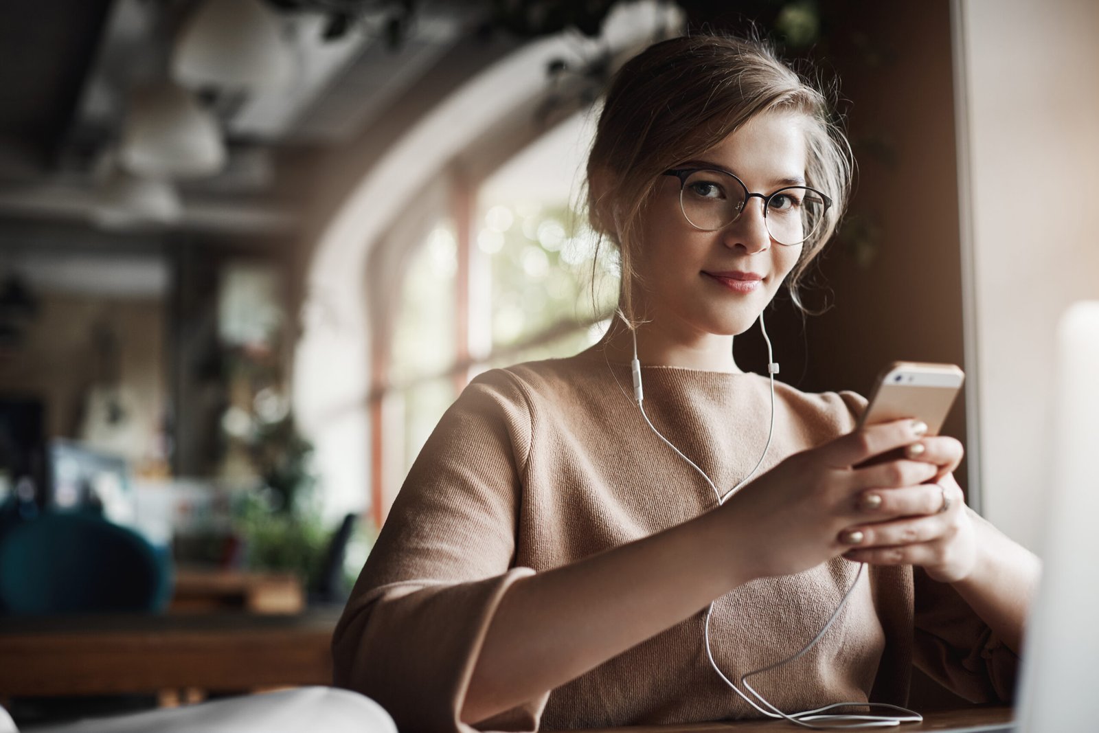 Indoor shot of good-looking stylish and happy caucasian female with fair hair in glasses, holding smartphone and wearing earbuds while watching video, distracting to look and smile at camera. Lifestyle, people and technology concept