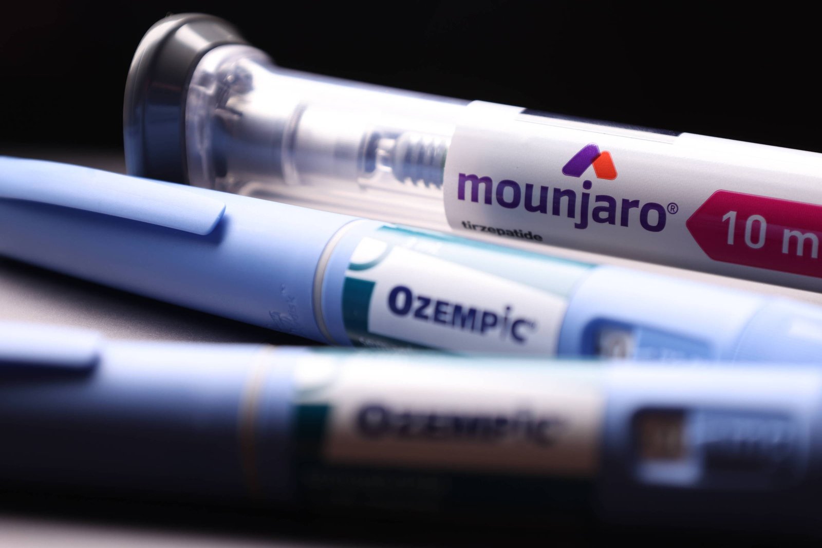 A imagem mostra três canetas de medicamento em primeiro plano. A caneta superior é transparente e contém o nome