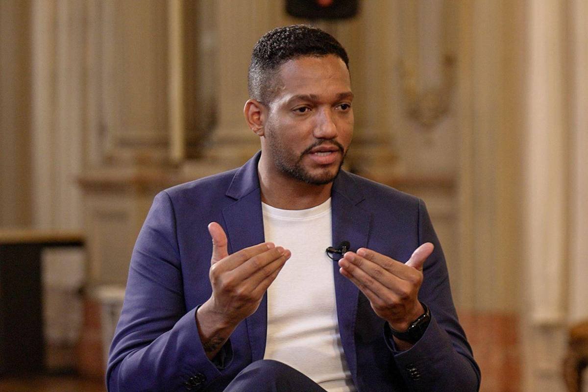 Homem de pele escura, cabelo curto e barba aparada, veste blazer azul e camiseta branca, gesticulando com as mãos enquanto fala em ambiente interno com decoração clássica.