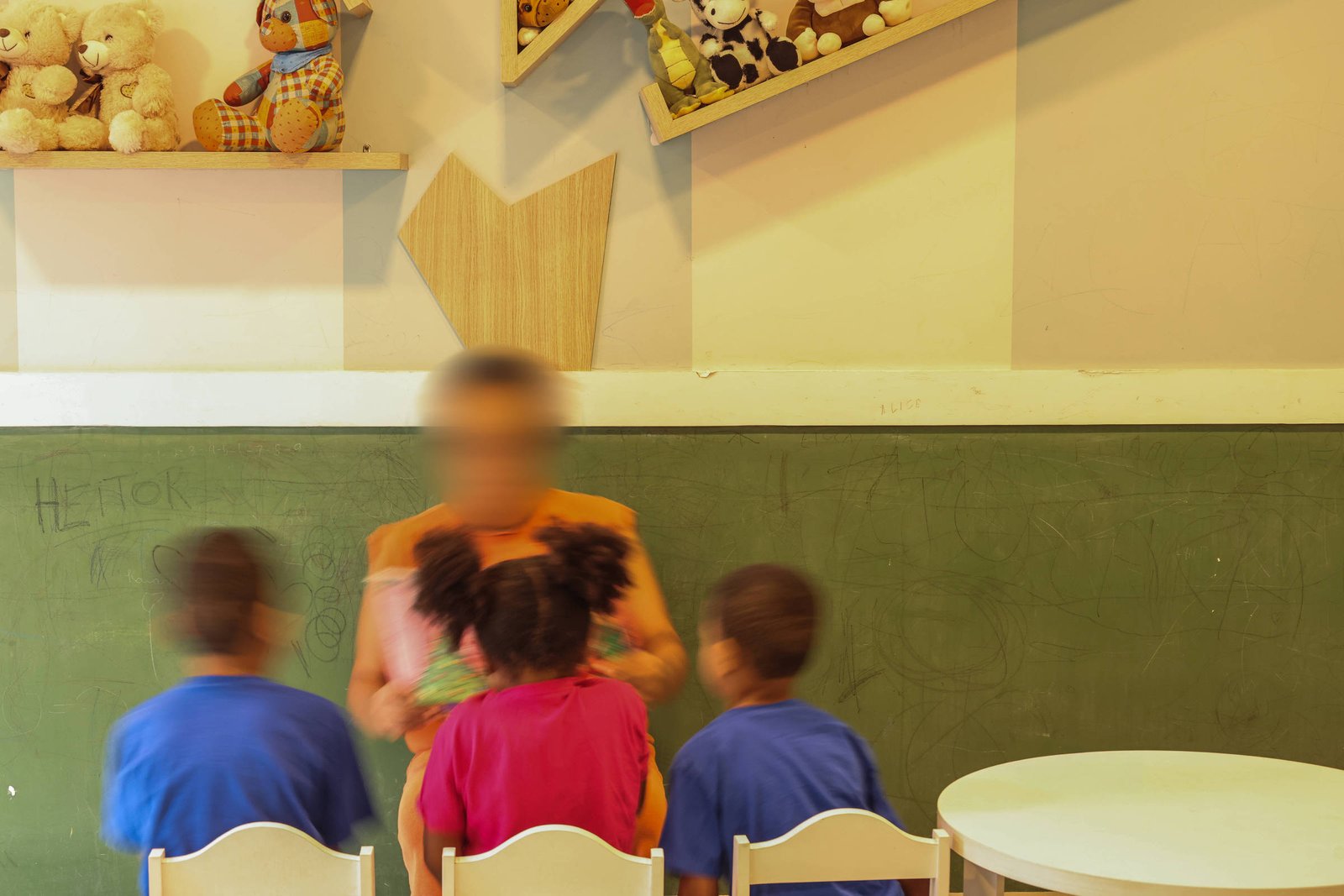 Três crianças sentadas de costas em cadeiras brancas diante de uma professora em pé, com quadro-negro verde ao fundo. Prateleiras com bichos de pelúcia decoram a parede clara.