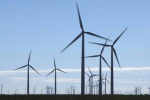 Diversas turbinas eólicas alinhadas em campo aberto sob céu azul claro com poucas nuvens. As pás das turbinas estão em movimento, indicando geração de energia eólica.