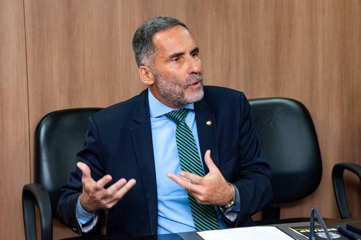 Homem de meia-idade com barba grisalha veste terno azul escuro, camisa azul clara e gravata verde listrada. Ele está sentado em cadeira preta em sala com parede de madeira, gesticulando com as mãos enquanto fala. Sobre a mesa à sua frente há documentos e um microfone.