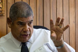 Homem de meia-idade com cabelo curto grisalho veste camisa branca e gravata preta, sentado em cadeira diante de parede de madeira, gesticulando com a mão direita levantada.