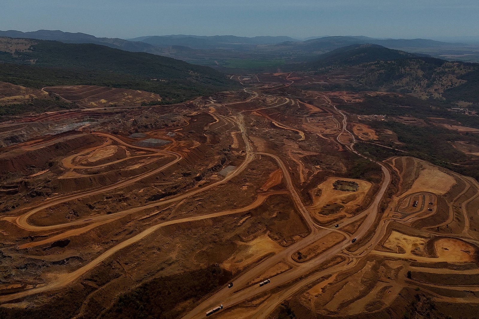 Mina a céu aberto com várias estradas de terra em ziguezague e áreas escavadas em diferentes níveis. Caminhões de mineração circulam pelas vias. Montanhas e vegetação ao fundo sob céu claro.