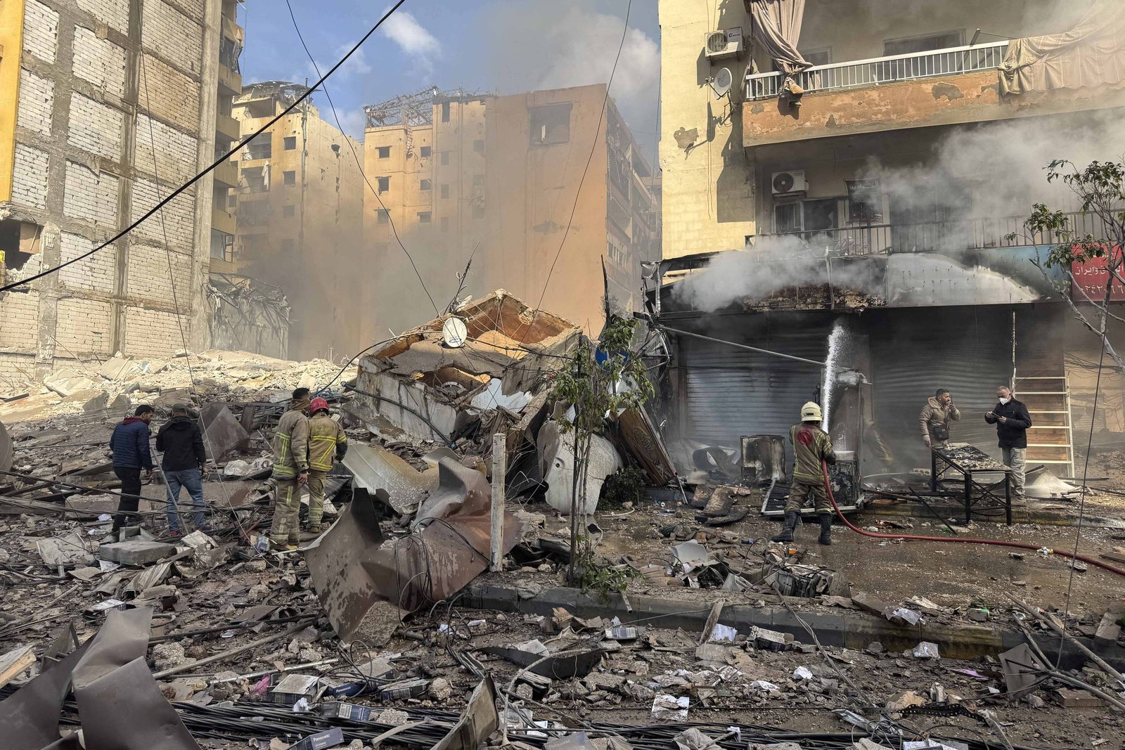 Equipes de resgate e civis caminham entre escombros e destroços de edifícios destruídos em rua urbana. Fumaça sai de uma loja com portas metálicas fechadas, enquanto prédios ao fundo mostram danos estruturais e janelas quebradas.