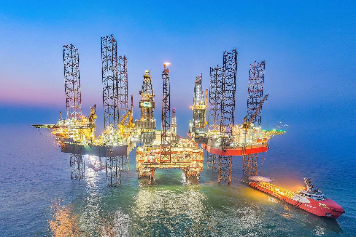 Cinco plataformas de petróleo elevadas sobre o mar, iluminadas com luzes amarelas, cercadas por água calma ao entardecer. Um navio de apoio vermelho está ancorado próximo à direita.