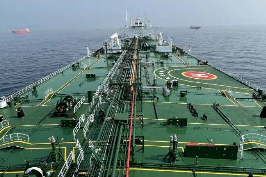Convés amplo de navio petroleiro verde com tubulações e equipamentos, incluindo heliponto marcado com H. Mar calmo ao redor e outros navios ao fundo.
