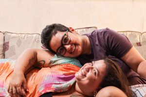 Duas pessoas deitadas em sofá com estofado claro e almofadas estampadas. Uma veste camiseta roxa e óculos, apoiando a cabeça sobre o ombro da outra, que usa blusa colorida laranja e rosa. Ambas sorriem para a câmera em ambiente interno.