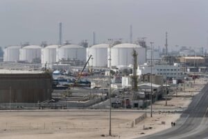 Vista ampla de um complexo industrial com vários tanques cilíndricos brancos de armazenamento de combustível ao fundo. Estruturas metálicas, guindastes e edifícios industriais ocupam o centro da imagem. Duas pessoas caminham pela estrada asfaltada à direita, que atravessa a área desértica e cercada por cercas.