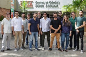Dez homens e uma mulher posam em fila na entrada do Centro de Ciências Sociais Aplicadas, com plantas ao redor e o nome do centro visível na fachada.