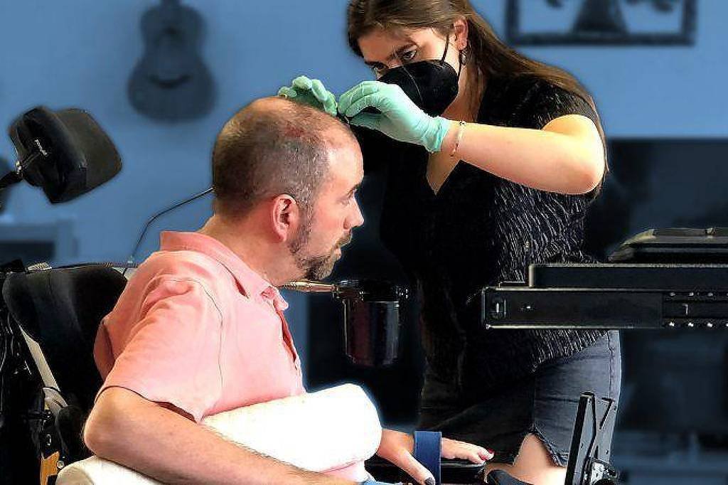 Mulher com máscara e luvas verdes examina a cabeça de homem sentado em cadeira de rodas em ambiente interno com instrumentos ao fundo.