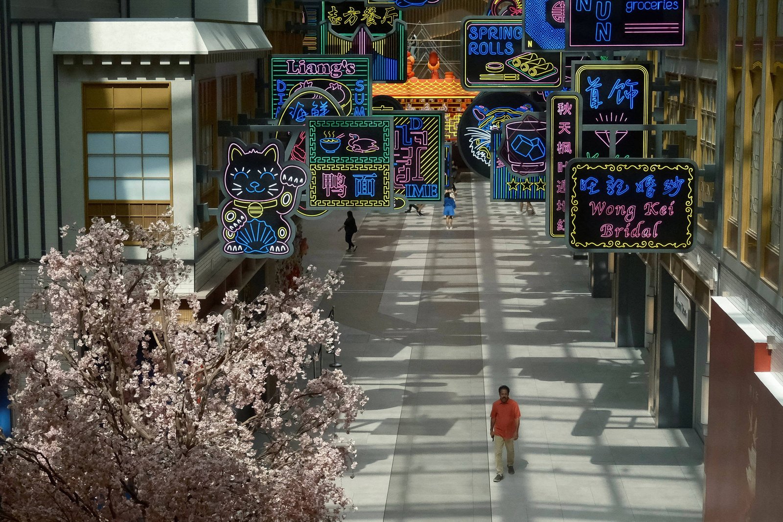 Corredor interno iluminado por luz natural com letreiros de néon coloridos pendurados no teto, exibindo nomes de lojas e símbolos em inglês e caracteres asiáticos. Árvore com flores cor-de-rosa à esquerda e poucas pessoas caminhando pelo espaço.