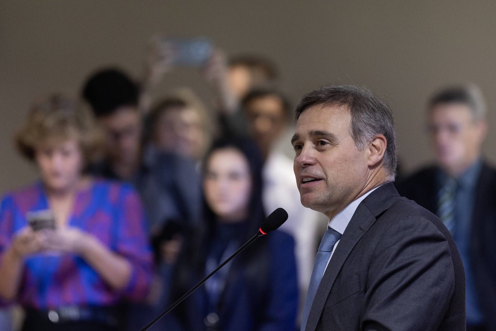 Homem de terno cinza e gravata azul fala ao microfone em ambiente interno. Ao fundo, grupo de pessoas desfocado, algumas olhando para celulares e outras observando o orador.