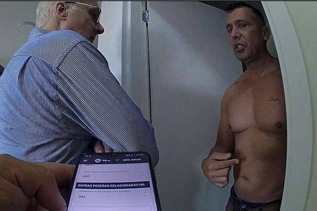 Homem sem camisa encostado em parede conversa com homem de camisa azul em ambiente interno. Celular com tela visível está sobre mesa próxima. O homem de camisa azul tem cabelos brancos e usa óculos, enquanto o homem sem camisa tem cabelos escuros e bem aparados.