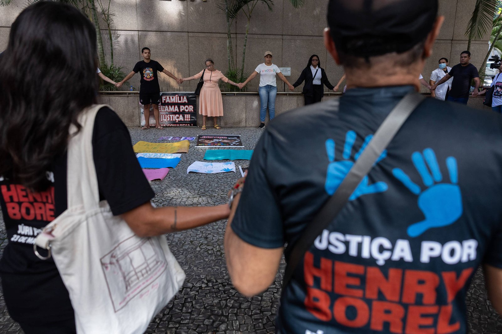 Grupo de pessoas de mãos dadas em protesto contra violência, com cartaz e faixas no chão. Dois manifestantes em primeiro plano usam camisetas com a frase