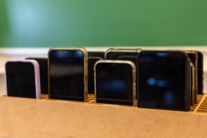 A imagem mostra um suporte de madeira com vários smartphones organizados. Os dispositivos têm telas pretas e estão dispostos em diferentes tamanhos e orientações. Ao fundo, há uma parede verde, sugerindo um ambiente de sala de aula ou escritório.