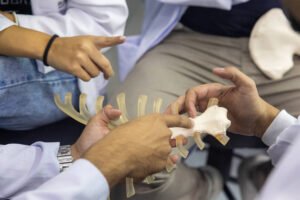 A imagem mostra um grupo de pessoas em um ambiente de aprendizado, onde uma pessoa está segurando um modelo de coluna vertebral e apontando para uma parte específica. Outra pessoa, com uma mão levantada, parece estar fazendo uma observação ou pergunta. As pessoas estão vestindo jalecos brancos, indicando que estão em um contexto acadêmico ou profissional relacionado à saúde.