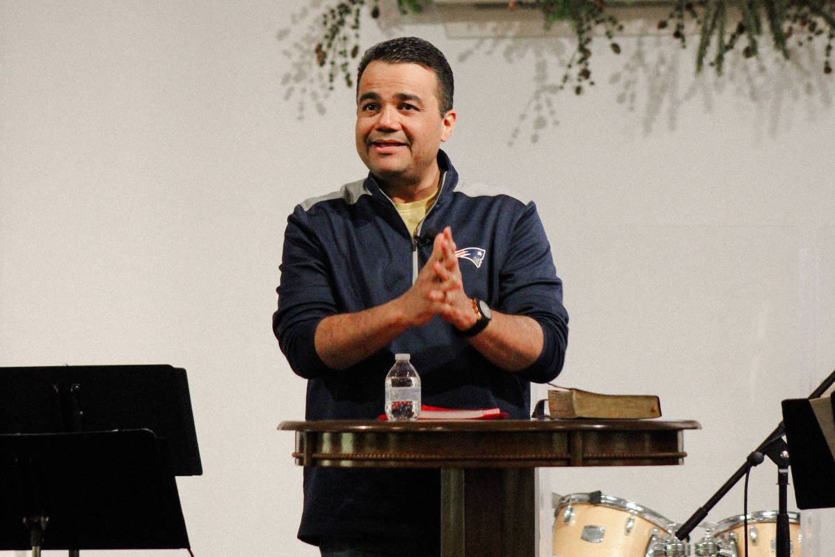 Homem de camiseta azul escura fala em púlpito de madeira com Bíblia e garrafa de água. Ao fundo, arranjo floral com flores brancas e vermelhas e bateria musical.