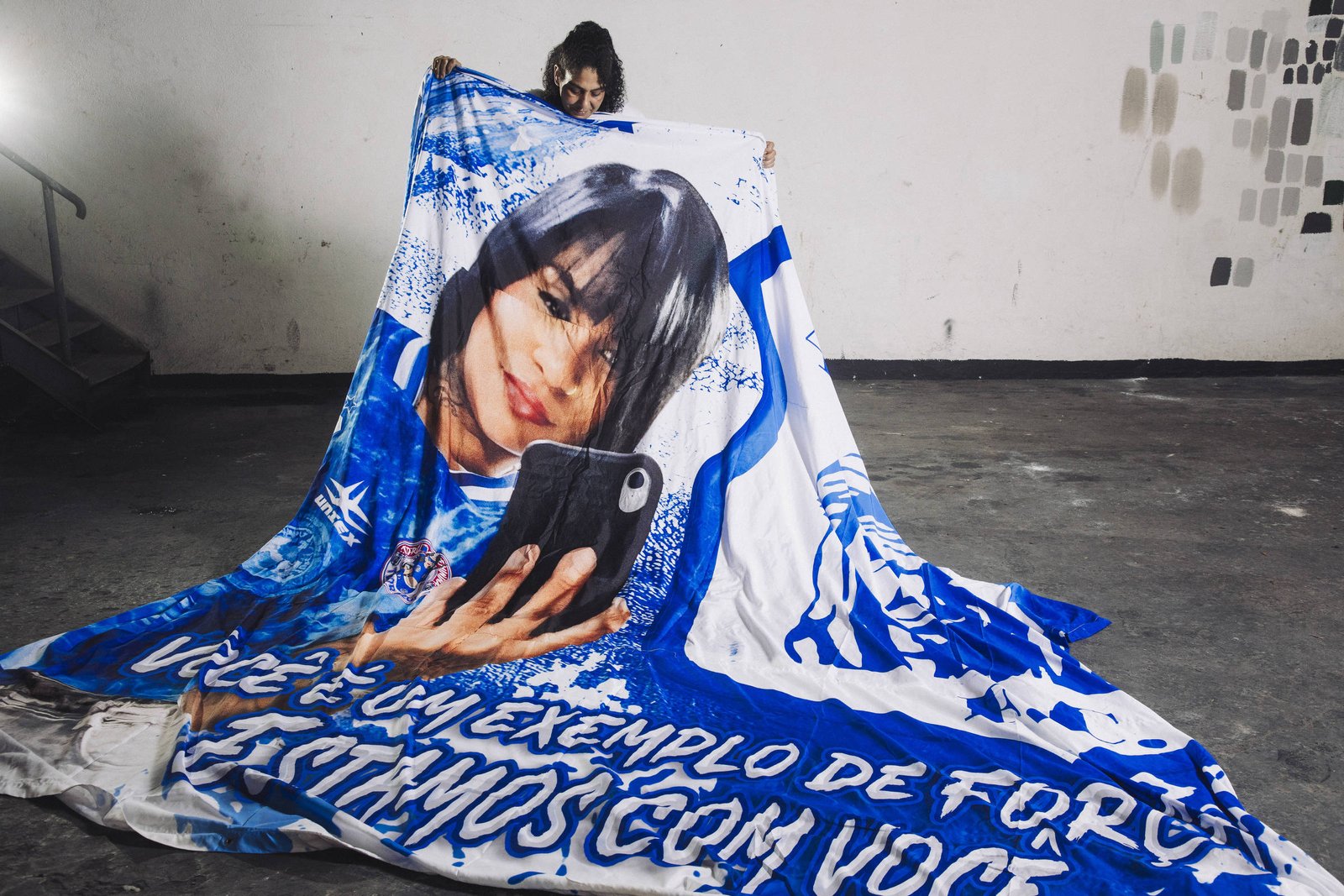 Pessoa em ambiente interno segura uma grande faixa azul e branca com a imagem de uma mulher tirando selfie e texto em português.