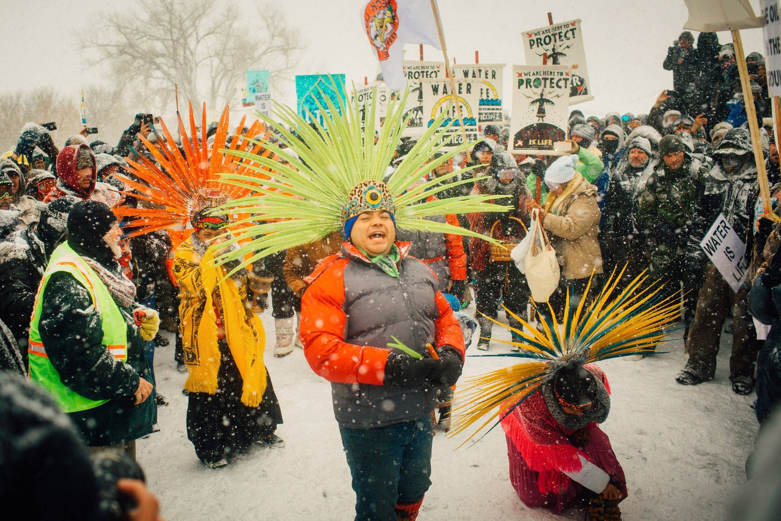 A imagem mostra um grupo de pessoas reunidas em um protesto durante uma nevasca. No centro, duas crianças vestindo roupas coloridas e adornos de penas dançam. Uma delas usa um casaco laranja e a outra um traje com penas verdes e amarelas. Ao fundo, há uma multidão de manifestantes segurando cartazes que dizem