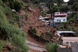 Deslizamento de terra cobre encosta e destrói várias casas em área residencial com vegetação ao redor. Terra e destroços bloqueiam rua abaixo.