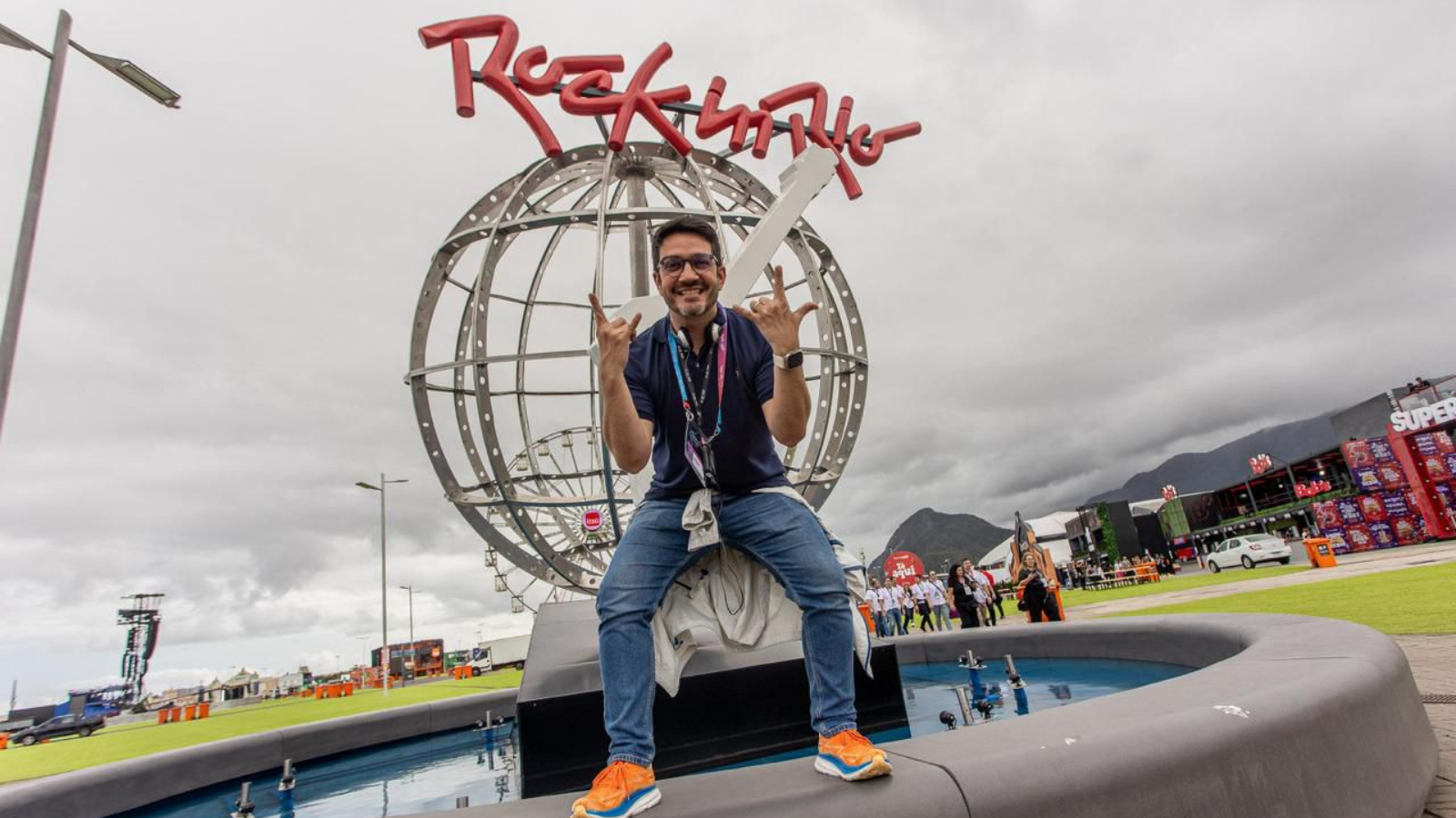 Marcelo Braga posando em frente à estrutura icônica do Rock in Rio, em ambiente externo de evento, reforçando conexão com grandes produções e experiências ao vivo.