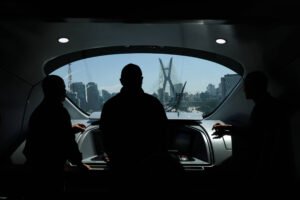 Três silhuetas de pessoas estão dentro de uma embarcação, olhando pela janela frontal para a Ponte Estaiada em São Paulo, com o horizonte urbano ao fundo em dia claro.